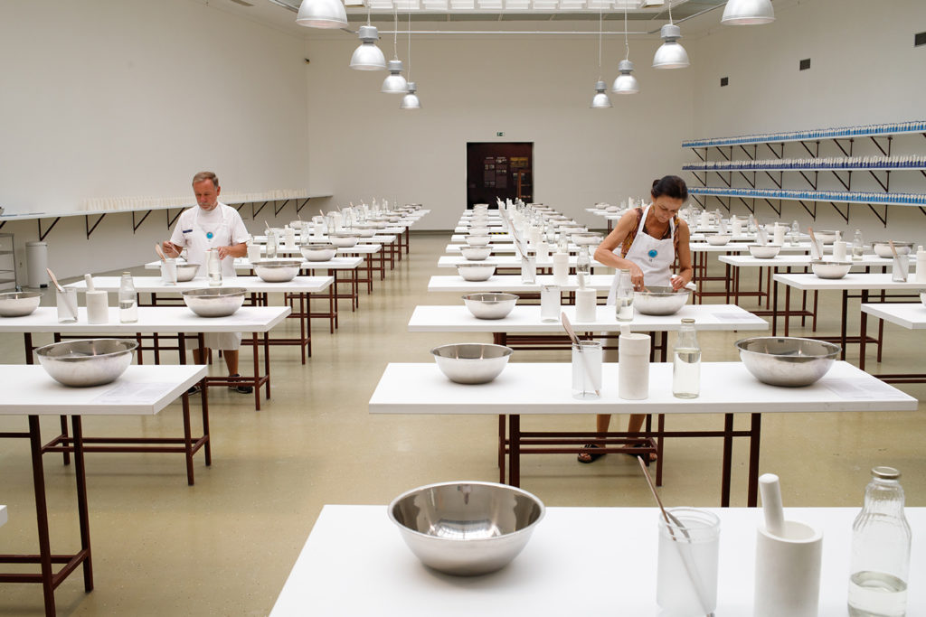 A room full of tables with mixing bowls, and other cooking equipment. Two people are at two different tables.