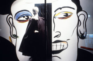 Mannequin in front of two faces painted on a wall.