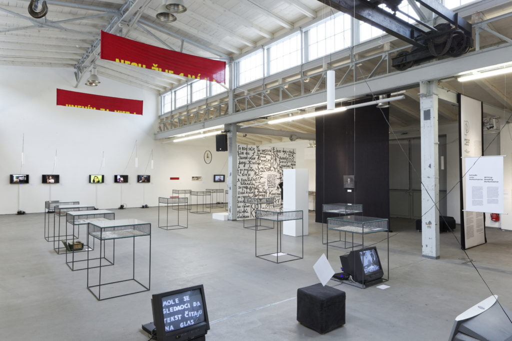 installation view of a gallery with TVs on the floor and vitrines with wiry bases