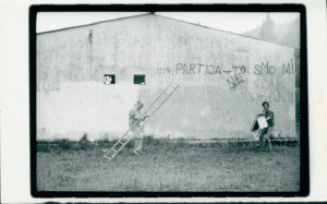 Black and white image of two people in front of a wall. The person on the left has a ladder.