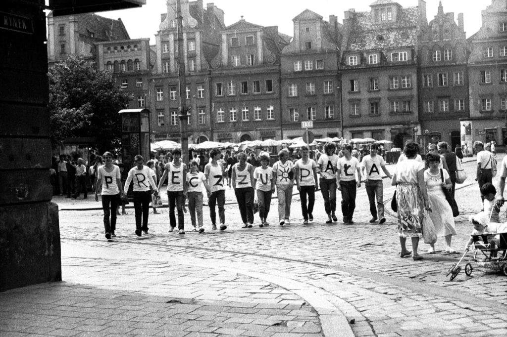 Black and white image of people in dark pants and light tops standing on a street in front of a row of buildings. Each person has a letter on their shirt.