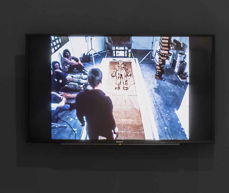 people sitting on the floor to the left of an installation featuring a lying human figure on the ground and one person in the foreground in black-view, many books piled up in the right side of the background
