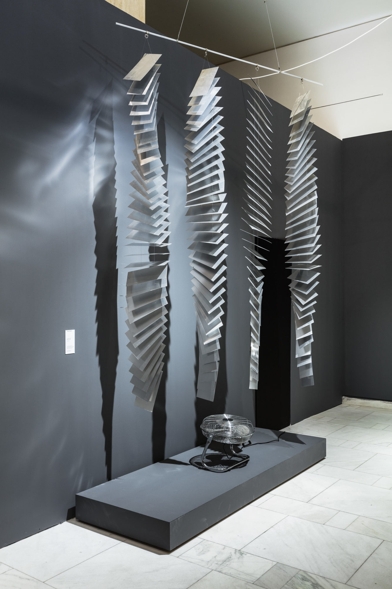 square metal plates stringed in four vertical garlands in front of a dark grey wall and with a fan placed under them