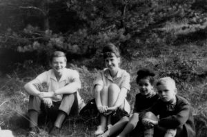 A photograph of the filmmaker as a child, accompanied by her mother, step father, and half-brother. They are sitting down in the grass together. The filmmaker has visibly darker skin than the rest of her family.
