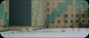 The exterior of a block of Soviet apartments, with light green and yellow geometric patterns on the exterior. There is snow in the yard, and a yellow ladder, as well as several large balls of snow