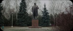 A view of a bronze statue of Lenin in the middle of a park, with two evergreen trees to either side.