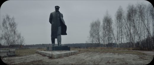 A view of a Lenin monument against a bleak field with bare trees to either side. The statue shows Lenin walking forward, his coat blown open by the wind.