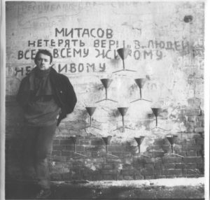 A black and white photo of the artist (a man with short dark hair wearing a dark jacket) standing by a brick wall with a series of 10 funnels attached to the wall
