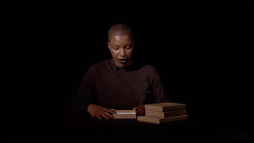 The artist, a black woman with very short hair, sitting at a table against a dark background. She has a box open in front of her and appears to be reading something inside the box.