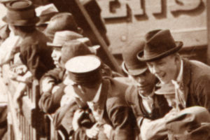A sepiatone photograph showing people in hats looking and laughing at something they see to the lower left.
