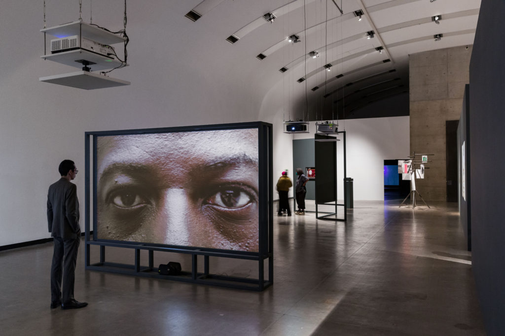 A view of the interior of an exhibition at the kunsthalle wien,with people observing video screens. In tthe foreground, a screen shows a Black man's eyes close-up, looking out from the screen.