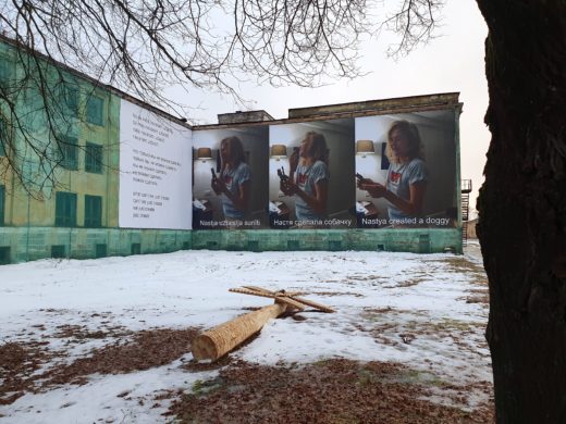 In the foreground, a palm tree made of roughly cut wood lies on snowy ground while n the background there are large text and photo panels installed on old buildings. One group of three panels shows a woman with a toy, with a caption in three languages
