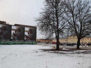 In a courtyard between buildings, there are two large trees and a sculpture of a palm tree that is lying on the ground. In the building in the background there are three large panels with images of a woman making a toy. The images are captions "Natastya created a doggy" in three languages.
