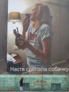 An image of a large scale photograph blown up and installed on the side of a building. The photo is of a woman with a toy dog. The image has text below it in Russian, reading, "Natastya created a doggy." There is a person at lower left adjusting the edge of the large installed photo.