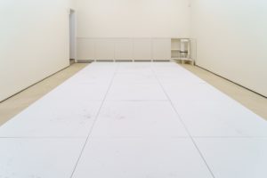 A white room with a grid of drawings laid out on the floor. The drawings are of dust and detritus and are done against a white background.