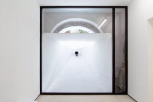 A wall-height window from a white room to the outside has a large white tent placed against it, allowing a person to place their head inside and look into the room from outside.
