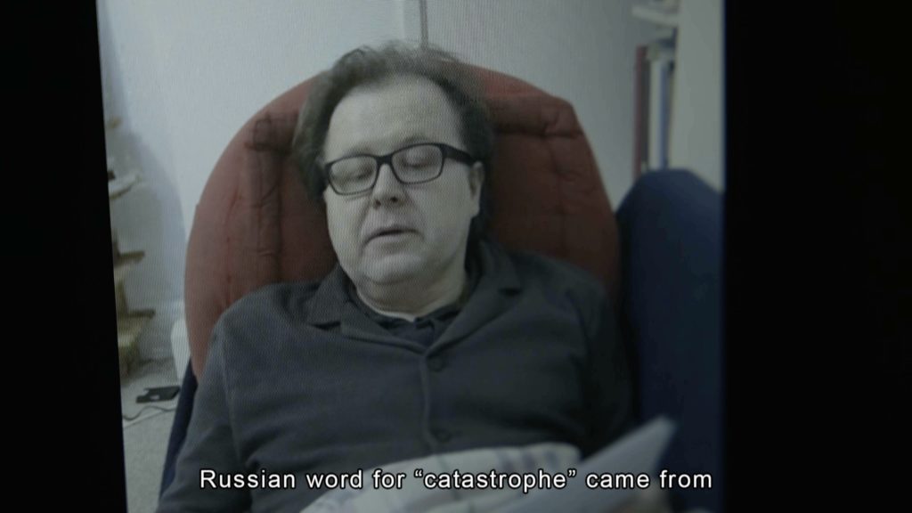 A man sitting on a chair and reading a text. The title on the bottom of the still reads "Russian word for 'catastrophe' came from."