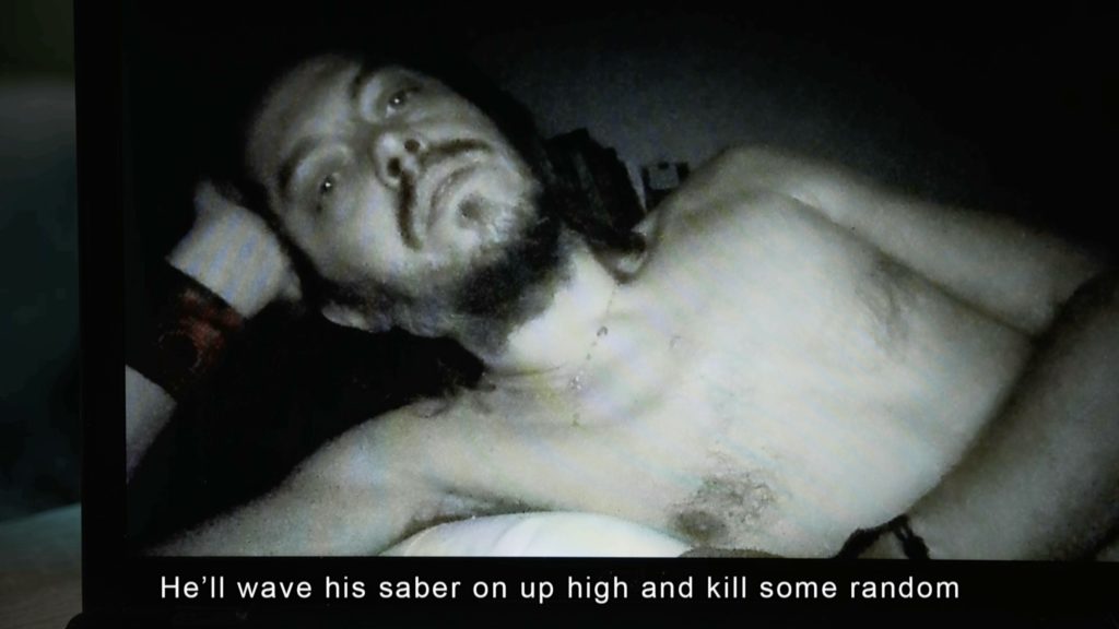 A man lying on the bed. The caption on the bottom of the still reads "He'll wave his saber on up high and kill some random."
