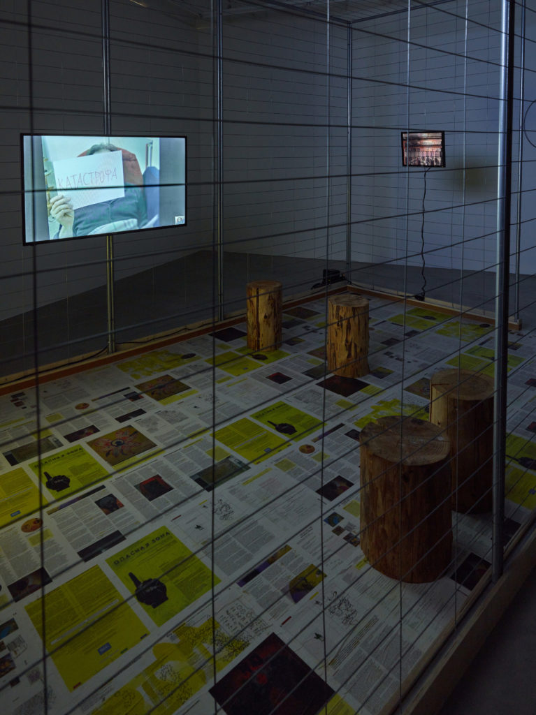 The Canary Archives installation comprising a large metal cage and TV screens. Wood logs used as seats. The floor is covered with newspaper pages. The image on one of the screens shows an artist holding a sign that reads "Catastrophe."