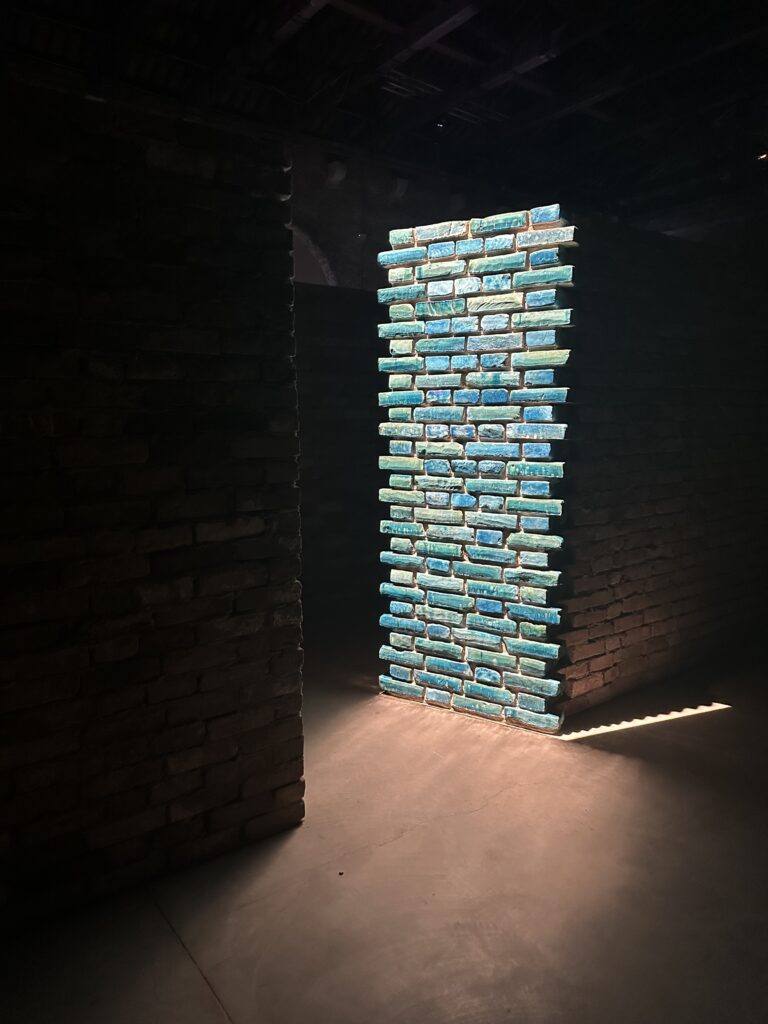 Within a labyrinth of brick walls, one end of a brick wall at a corner of the labyrinth is brightly lit, with blue ceramic bricks illuminated prominently.