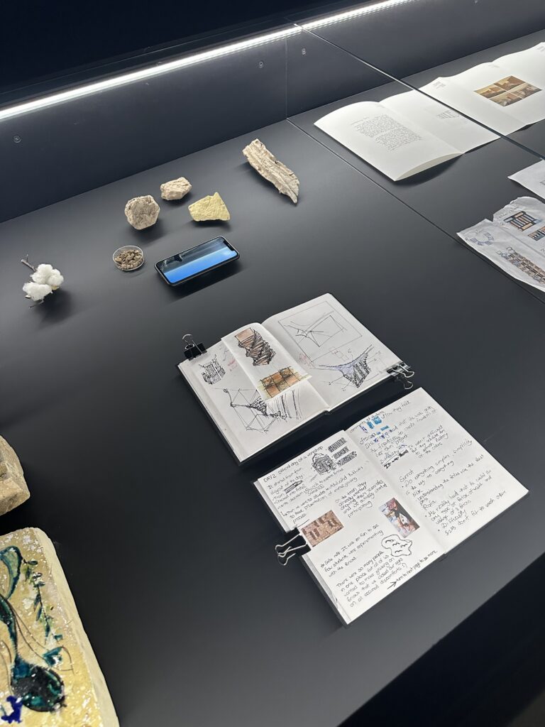A collection of objects laid out in a vitrine with a dark grey background. The objects include a cell phone, stones, and sketchbooks with clips holding open the pages.