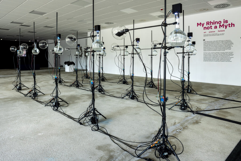 Exhibition space with three rows of identical objects, each consisting of a stand holding two glass objects. The objects are connected with cables.