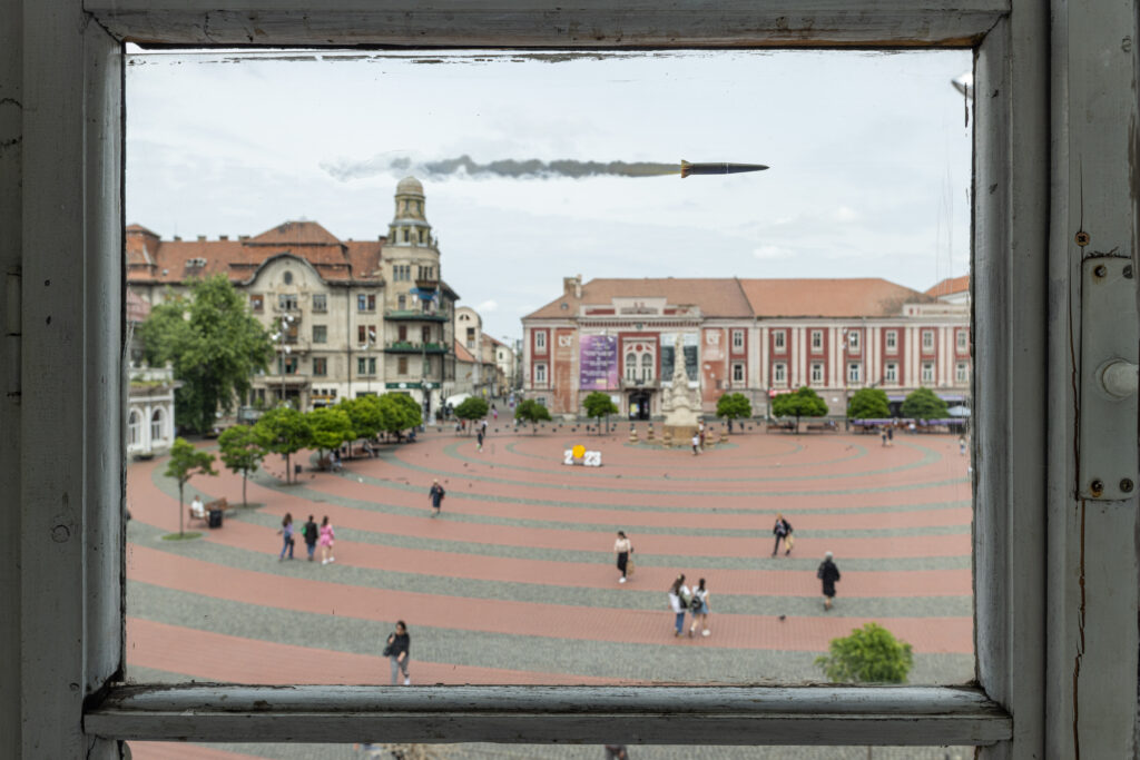 Image of a city square seen through a window, with a missile flying across the sky.