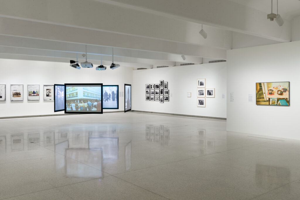 An installation view of a museum, with white walls and a glossed gray floor. At center left, four screens hang suspended from the ceiling in a square configuration. At right, there are several configurations of framed photographs hanging on the walls. Not all the images are legible, but one of them (at center right) is Sanja Ivekovic's Triangle, which features three black and white photos arranged vertically at left and one at center on the right, showing the artist on a balcony reading.