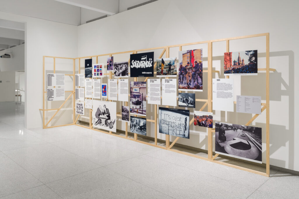 A view of a museum exhibition space, with an informational display constructed of bare wood scaffolding, with images and text blocks printed and mounted on the scaffolding. The textual blocks are divided into chronological periods (for example, 1975-1980, 1981-1986, Revolutions of 1989, and 1991). Among the photographs that are mounted on the scaffolding, one of them is an image of the "Solidarnosc" logo, and another shows a graffiti that reads "It's Over! Czechs are free!"