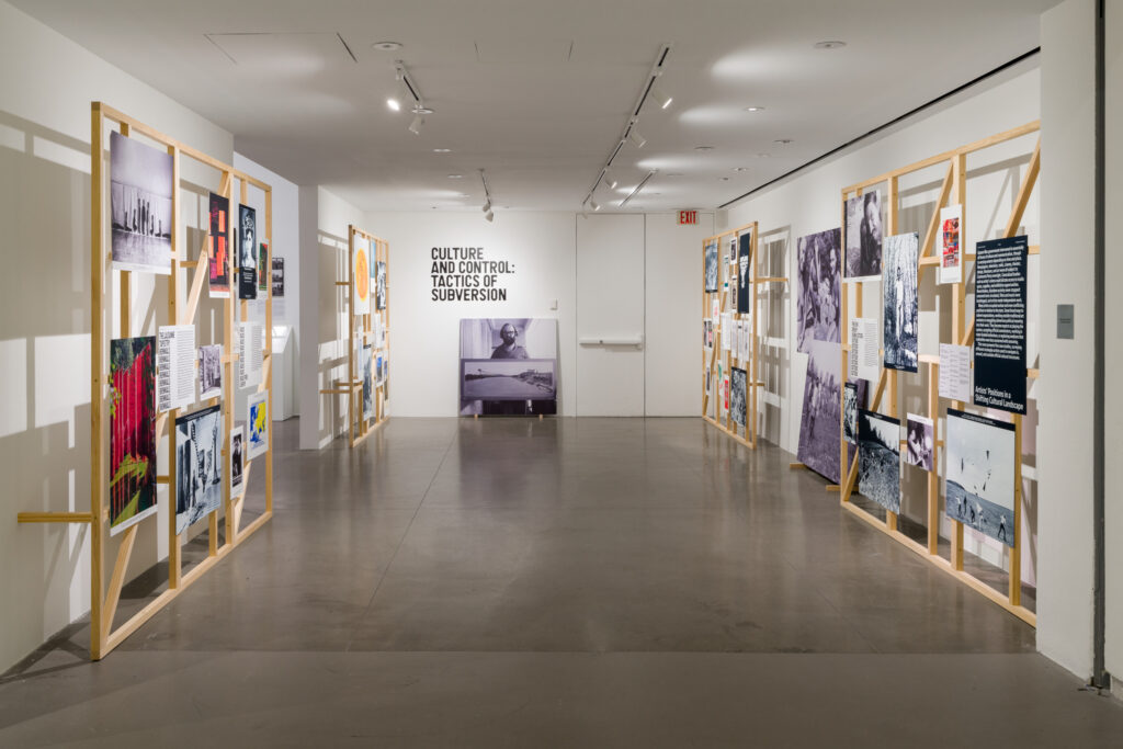 A view of a museum exhibition space with four wooden scaffolding structures containing informational text and photographs, arranged on walls to either side of the photograph's view. At center, at the far end of a rectangular space, we see a white wall with the exhibition section title "Culture and Control: Tactics of Subversion" visible. Below that is a black and white photograph of a man holding a landscape photograph.