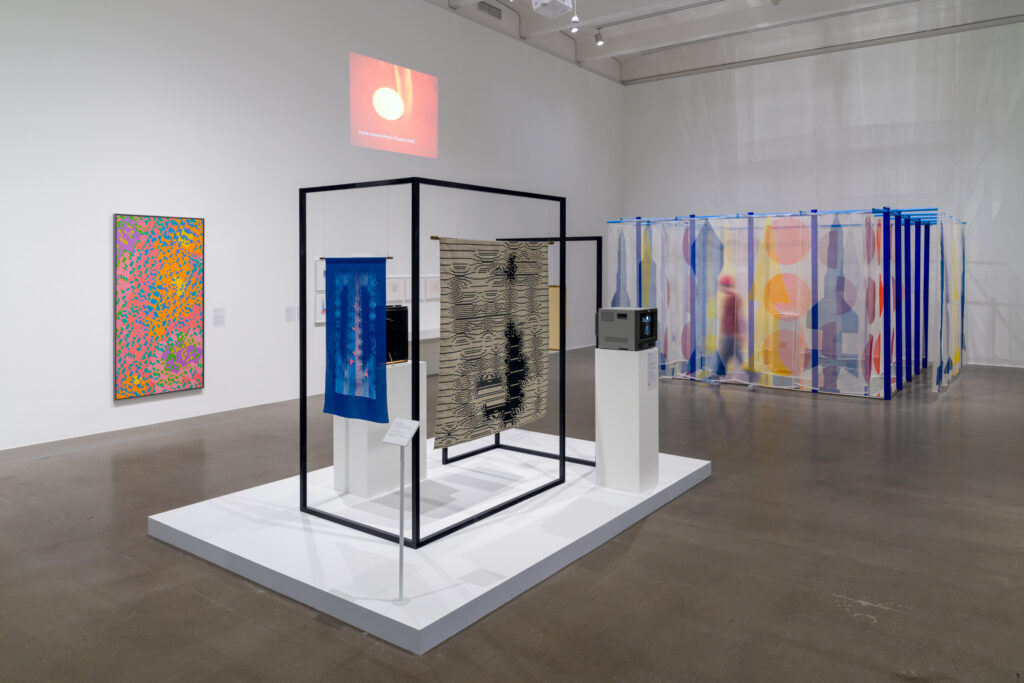 An installation view of a museum with white walls and a glossy gray floor. We are looking into a broad corner of the museum, and in the space we see three-dimensional displays of textile works, some mounted on rectangular frames. The works are brightly colored, with a combination of organic, wavy patterns and (in the work furthest from us) silhouettes that recall rockets or spaceships. There is also a video monitor mounted on the raised floor section closest to us.
