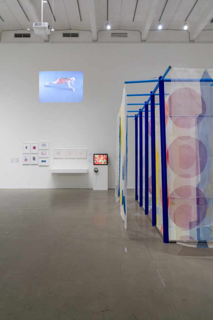 A view of a museum exhibition. At right there is a structure with blue rods and partially transparent textiles hanging from the structure. The textiles have simple geometric shapes (circles) on them. At left, we can see a video monitor on a pedestal, and a video projected on the wall above it as well. The video seems to depicts a woman against a blue background. There are some framed works on the wall at lower right.