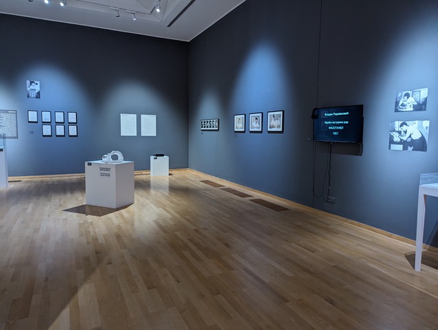 Exhibition view including a series of photographs and images on the wall, a television screen, and several display boxes on the ground.