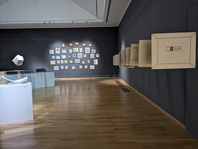 Exhibition view with a series of objects hanging form a ceiling in a row on the one side, display boxes on the ground and an arrangement of framed images on the wall in the back of the room.